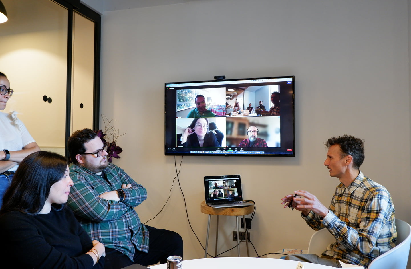 An Inviqa expert wearing a checkered shirt talks and gestures with his hands to people in a meeting room and remote people, visible on a wall-mounted TV
