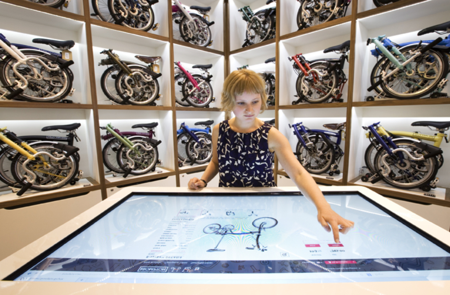 A sales assistant with cropped blond hair uses a giant touch screen to configure a Brompton bike, against a backdrop of folded bikes